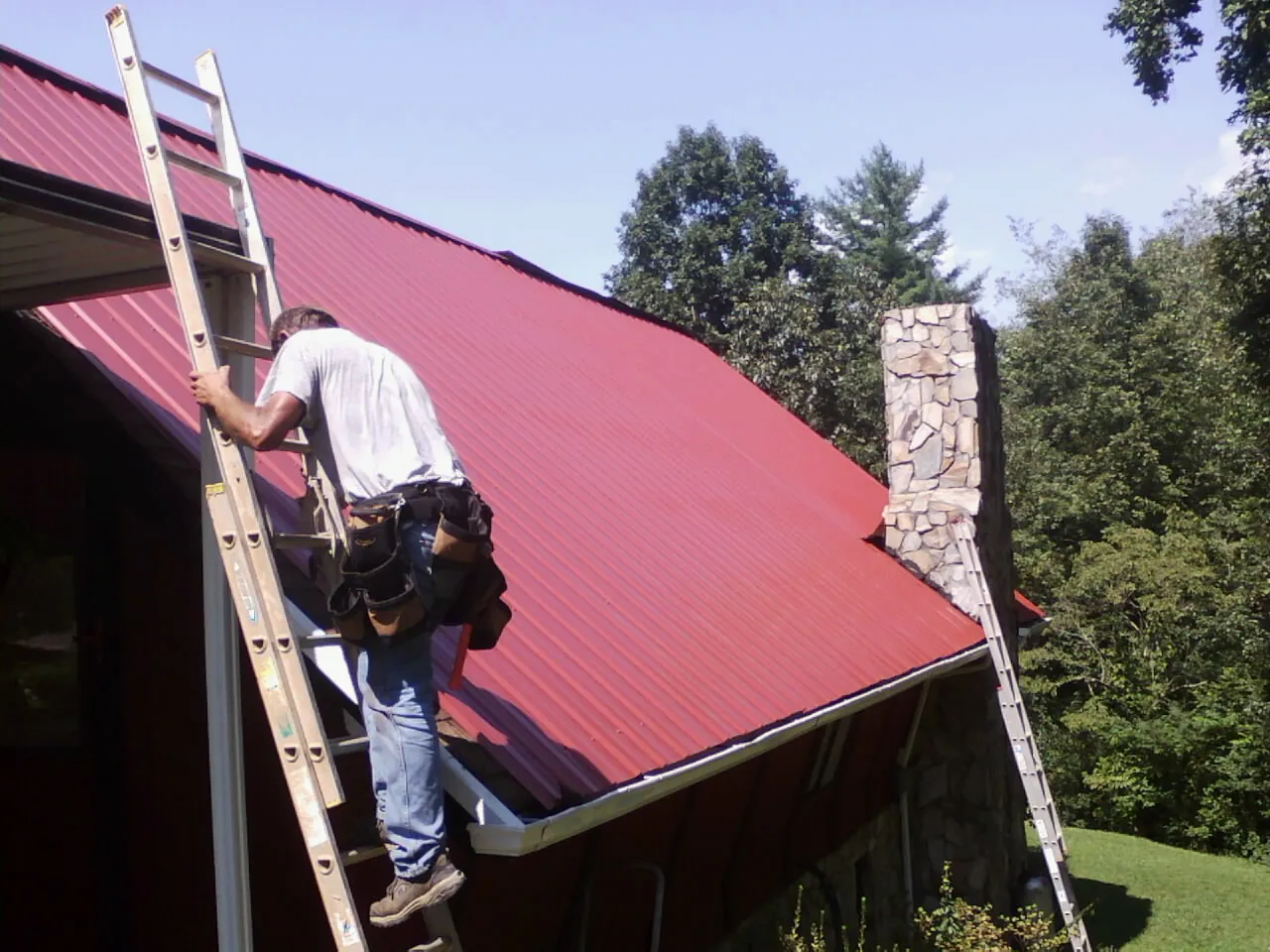 Roofer on a ladder beside a red metal roof and stone chimney — Soffit & Fascia Repair by Roofing Brothers in Central